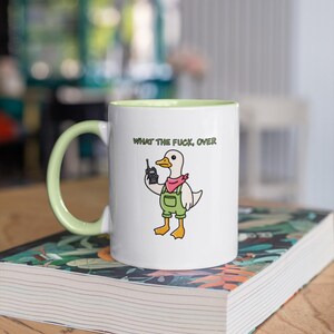 May include: White ceramic mug with a light green handle and interior. The mug features a cartoon duck wearing overalls and a pink bandana, holding a walkie-talkie. Text above the duck reads "WHAT THE FUCK, OVER."