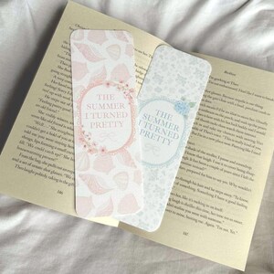 Op de afbeelding: Twee decoratieve bladwijzers met de tekst "The Summer I Turned Pretty". E&eacute;n bladwijzer heeft een roze schelpenpatroon en bloemaccenten, terwijl de andere een blauw bloemenontwerp heeft. De bladwijzers liggen op de pagina's van een boek.