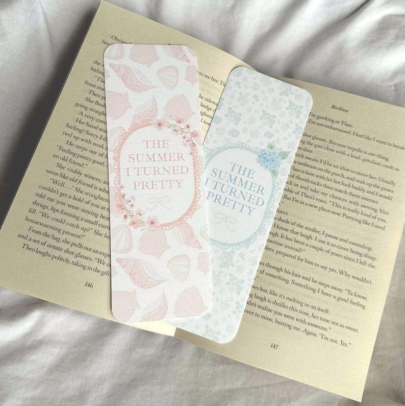 Op de afbeelding: Twee decoratieve bladwijzers met de tekst "The Summer I Turned Pretty". E&eacute;n bladwijzer heeft een roze schelpenpatroon en bloemaccenten, terwijl de andere een blauw bloemenontwerp heeft. De bladwijzers liggen op de pagina's van een boek.