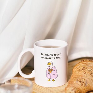 May include: White ceramic mug with the text "Bestie, I'm about to crash TF out." and a cartoon duck wearing a pink robe. A croissant sits on a wooden board next to the mug.