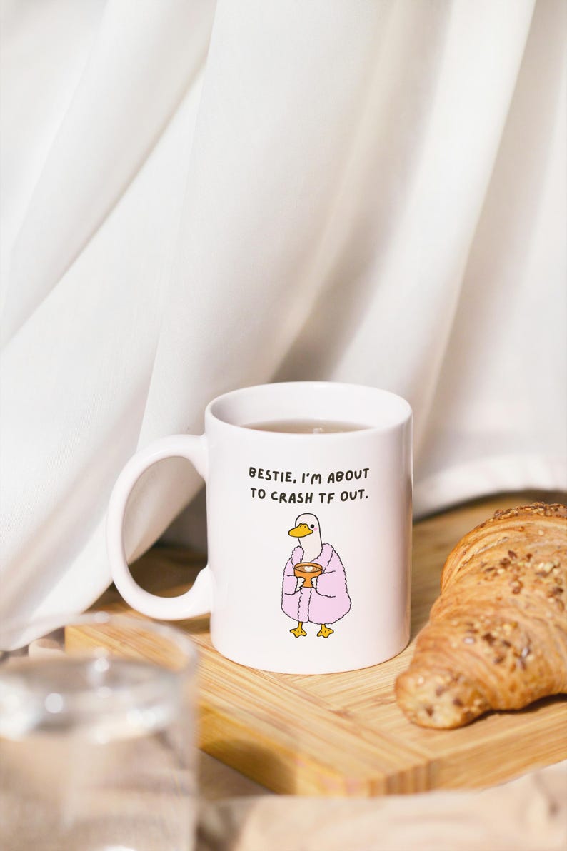 May include: White ceramic mug with the text "Bestie, I'm about to crash TF out." and a cartoon duck wearing a pink robe. A croissant sits on a wooden board next to the mug.