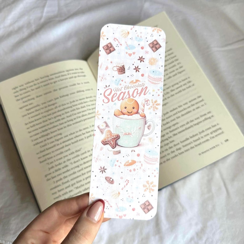 K&ouml;nnte beinhalten: Ein dekorativer Lesezeichen mit Winterthema. Das Lesezeichen zeigt einen Lebkuchenmann in einer Tasse mit dem Schriftzug "Hot Chocolate Season". Der Hintergrund ist wei&szlig; mit Illustrationen von Schneeflocken, Keksen und Schokolade.