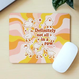 May include: A rectangular mouse mat with a retro design featuring cartoon ducks, stars, and the text "Definitely not all in a row." The background has a wavy pattern in shades of yellow, orange, and pink. A white computer mouse is on the right.