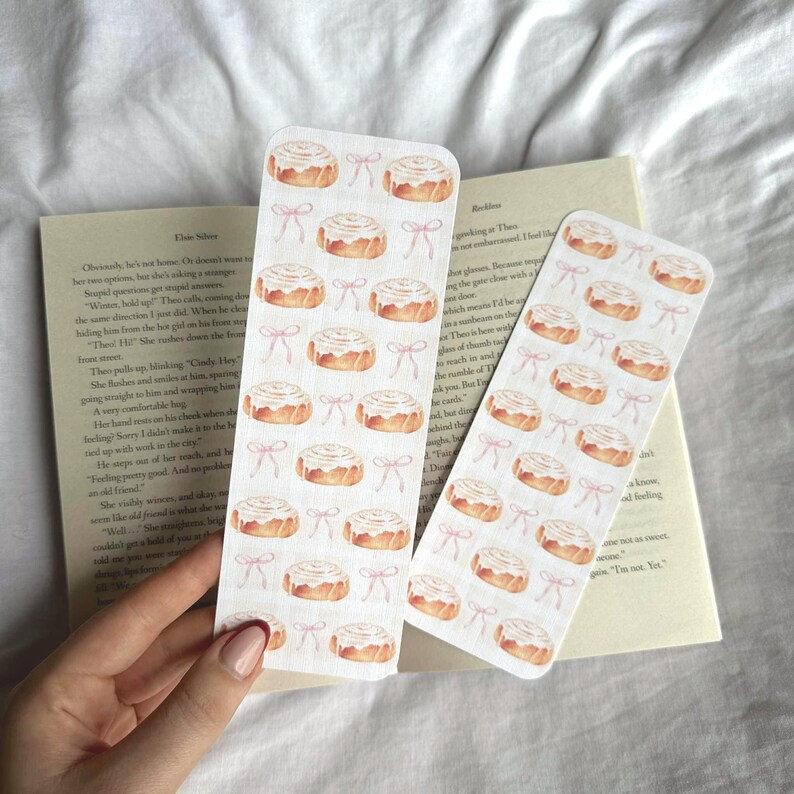 Pode incluir: Dois marcadores retangulares com um padr&atilde;o repetido de rolinhos de canela em aquarela e la&ccedil;os rosa. Os marcadores est&atilde;o em um livro aberto. O fundo &eacute; uma superf&iacute;cie branca texturizada.