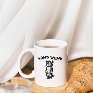 May include: White ceramic mug with the text "WOMP WOMP" and a black and white illustration of a standing kitten. A croissant sits on a wooden board next to the mug.