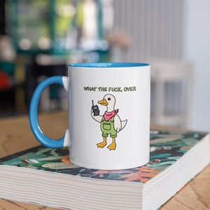 May include: White ceramic mug with a blue interior and handle. The mug features a cartoon duck wearing overalls and a pink scarf, holding a phone. The text "WHAT THE FUCK, OVER" is printed above the duck.