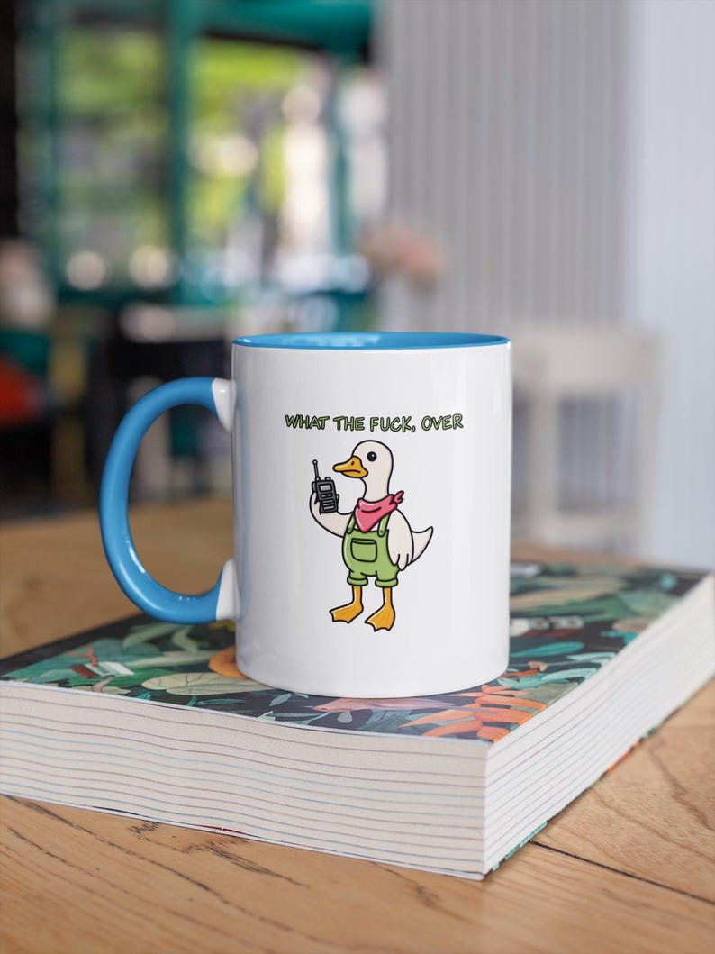 May include: White ceramic mug with a blue interior and handle. The mug features a cartoon duck wearing overalls and a pink scarf, holding a phone. The text "WHAT THE FUCK, OVER" is printed above the duck.