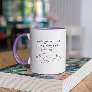 May include: White ceramic mug with a purple interior and handle. The mug features the text "undiagnosed but something defo ain't right" and a cartoon illustration of a goose. The mug is resting on a book.
