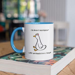 Puede incluir: Taza de cer&aacute;mica blanca con interior y asa azules. La taza presenta un pato de dibujos animados con un sombrero, con el texto "I'M BUILT DIFFERENT LIKE INCORRECTLY I THINK". La taza est&aacute; sobre un libro.