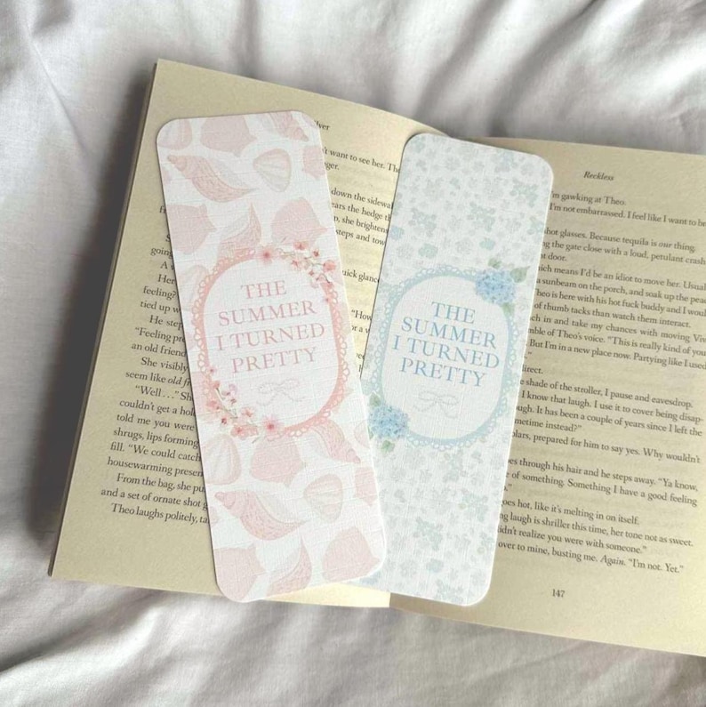 Op de afbeelding: Twee decoratieve bladwijzers met de tekst "THE SUMMER I TURNED PRETTY". E&eacute;n bladwijzer heeft een roze bloemenpatroon en de andere een blauw bloemenpatroon. Beide liggen op een open boek.
