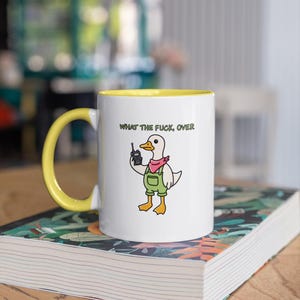 May include: White ceramic mug with a yellow interior and handle. The mug features a cartoon duck wearing overalls and a pink bandana, holding a retro cell phone. The text "WHAT THE FUCK, OVER" is printed above the duck.