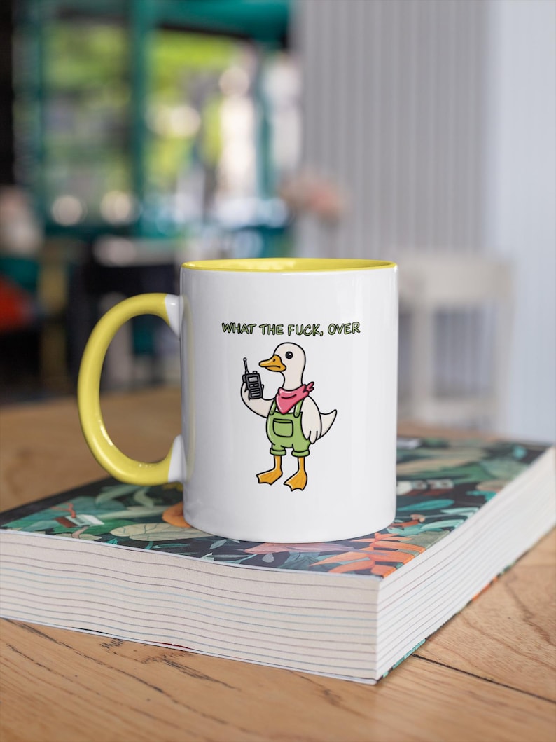 May include: White ceramic mug with a yellow interior and handle. The mug features a cartoon duck wearing overalls and a pink bandana, holding a retro cell phone. The text "WHAT THE FUCK, OVER" is printed above the duck.
