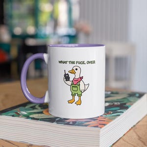 May include: White ceramic mug with a purple interior and handle. The mug features a cartoon duck wearing overalls and a pink scarf, holding a walkie-talkie. The text "WHAT THE FUCK, OVER" is printed above the duck.