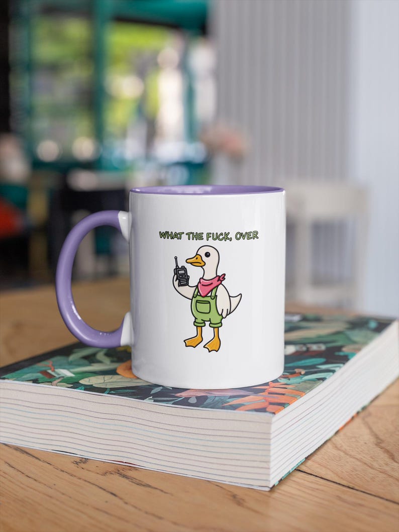 May include: White ceramic mug with a purple interior and handle. The mug features a cartoon duck wearing overalls and a pink scarf, holding a walkie-talkie. The text "WHAT THE FUCK, OVER" is printed above the duck.
