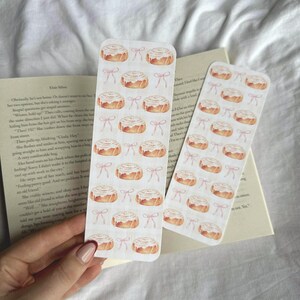 Pode incluir: Dois marcadores retangulares com um padr&atilde;o repetido de rolinhos de canela em aquarela e la&ccedil;os rosa. Os marcadores est&atilde;o em um fundo branco com um livro e uma m&atilde;o segurando um dos marcadores.