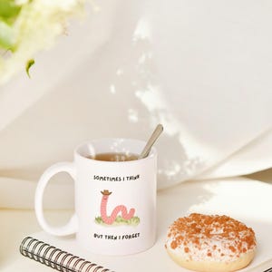 May include: White ceramic mug with the text "SOMETIMES I THINK BUT THEN I FORGET" and a cartoon worm wearing a cowboy hat. The mug contains tea and a spoon. A donut and a spiral notebook are also in the image.