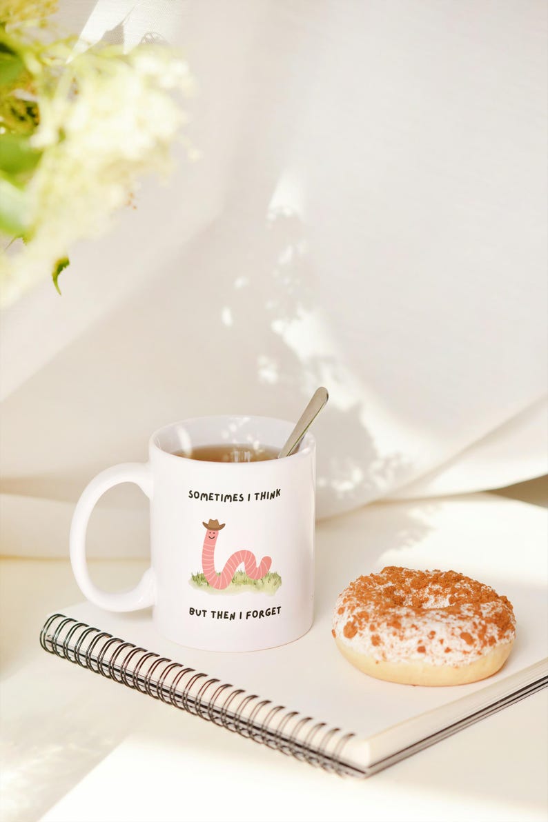 May include: White ceramic mug with the text "SOMETIMES I THINK BUT THEN I FORGET" and a cartoon worm wearing a cowboy hat. The mug contains tea and a spoon. A donut and a spiral notebook are also in the image.