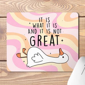 May include: A pink and orange mouse mat with a white goose lying on its back. The mouse mat has a wavy pattern and the text "It is what it is and it is not great."