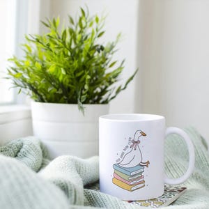 May include: White ceramic mug featuring a cartoon goose sitting on a stack of books. The mug has the text "stay home, read club". A green plant in a white pot sits next to the mug on a light green blanket.