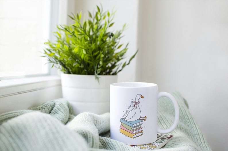 May include: White ceramic mug featuring a cartoon goose sitting on a stack of books. The mug has the text "stay home, read club". A green plant in a white pot sits next to the mug on a light green blanket.