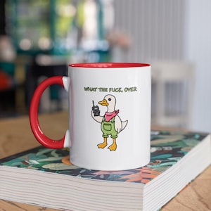 May include: White ceramic mug with a red interior and handle. The mug features a cartoon duck wearing overalls and a pink bandana, holding a walkie-talkie. The text "WHAT THE FUCK, OVER" is printed above the duck.