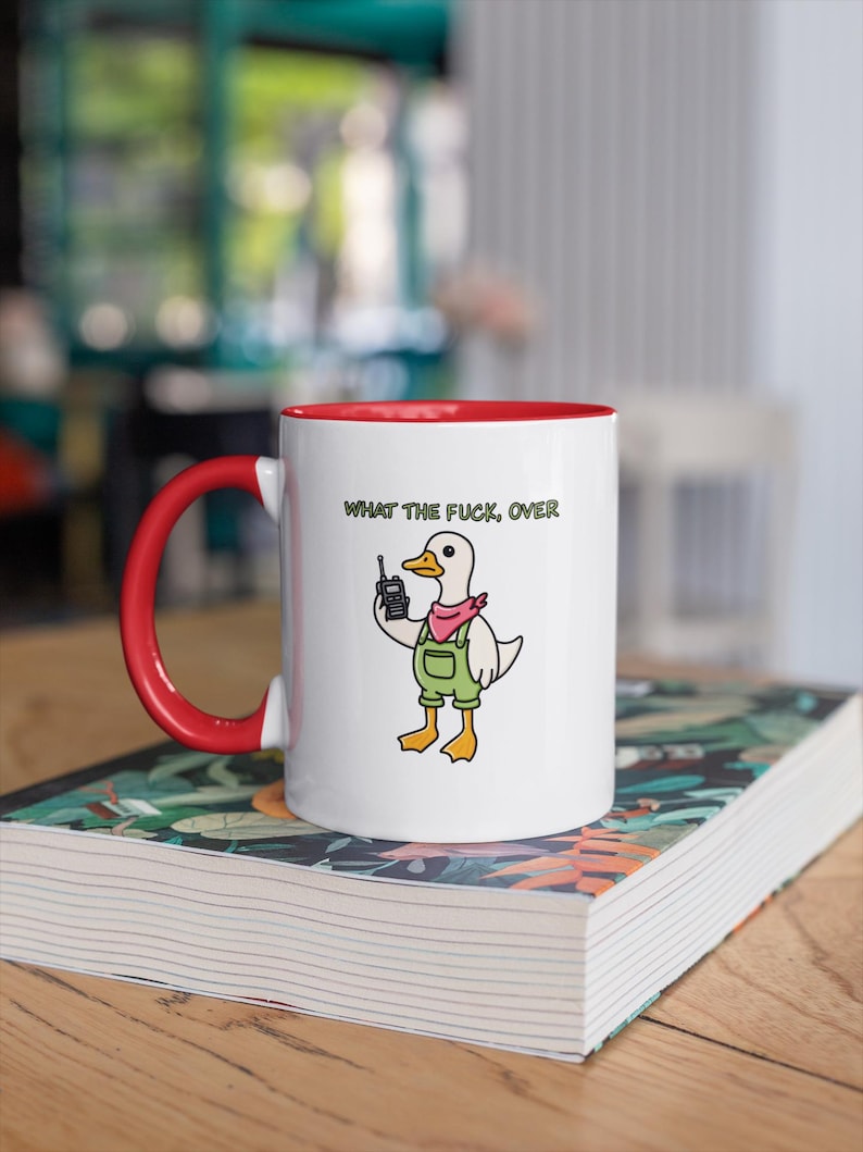 May include: White ceramic mug with a red interior and handle. The mug features a cartoon duck wearing overalls and a pink bandana, holding a walkie-talkie. The text "WHAT THE FUCK, OVER" is printed above the duck.