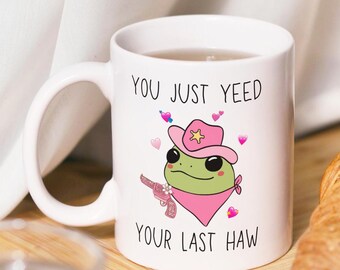 May include: White ceramic mug with a cartoon frog wearing a pink cowboy hat and bandana. The mug has the text "You just yeed your last haw" printed on it.
