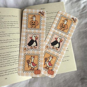 Op de afbeelding: Twee Halloween-thema bladwijzers met een lichtoranje ruitpatroon. Elke bladwijzer heeft stempelachtige illustraties van eenden in kostuums, waaronder een pompoen, vampier en duivel. Eikels en pompoenen zijn ook afgebeeld.