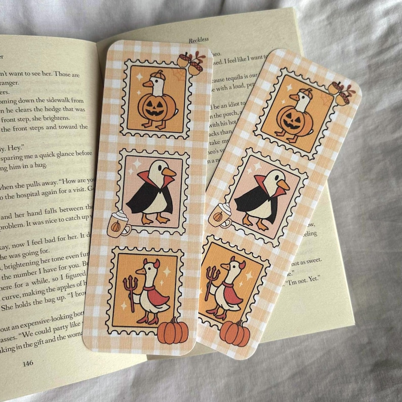 Op de afbeelding: Twee Halloween-thema bladwijzers met een lichtoranje ruitpatroon. Elke bladwijzer heeft stempelachtige illustraties van eenden in kostuums, waaronder een pompoen, vampier en duivel. Eikels en pompoenen zijn ook afgebeeld.
