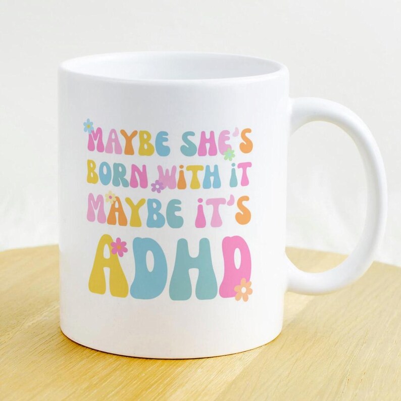 May include: White ceramic mug with the text "MAYBE SHE'S BORN WITH IT MAYBE IT'S ADHD" in colorful, retro-style lettering. The mug has a curved handle and is decorated with small flower graphics.