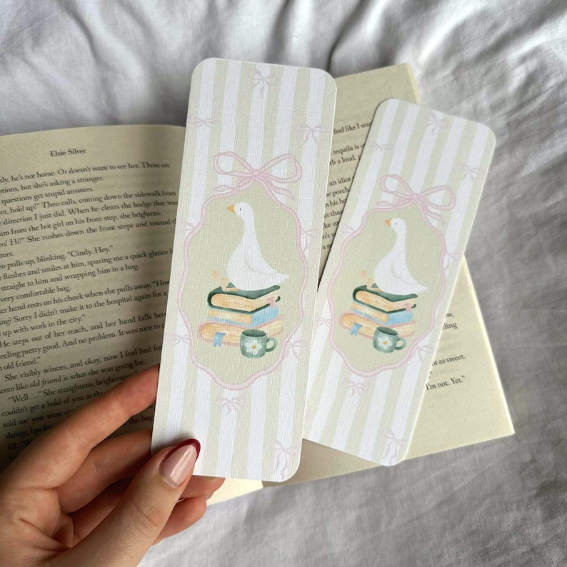 May include: Two decorative bookmarks with a whimsical design. Each bookmark features a white goose atop a stack of books, a teacup, and a pink bow. The background has vertical stripes and small bow accents.