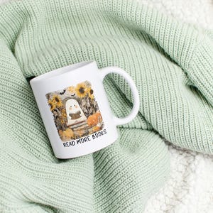 May include: White ceramic mug with a wraparound illustration of a ghost reading a book in a pumpkin patch, with sunflowers and the text "READ MORE BOOKS". The mug is on a light green knitted blanket.