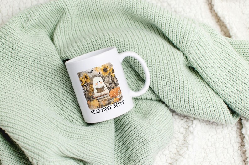 May include: White ceramic mug with a wraparound illustration of a ghost reading a book in a pumpkin patch, with sunflowers and the text "READ MORE BOOKS". The mug is on a light green knitted blanket.