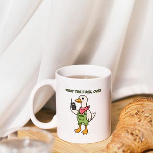 May include: White ceramic mug featuring a cartoon duck wearing overalls and a red bandana, holding a walkie-talkie. The mug has the text "WHAT THE FUCK, OVER" printed on it. A croissant sits on a wooden board.