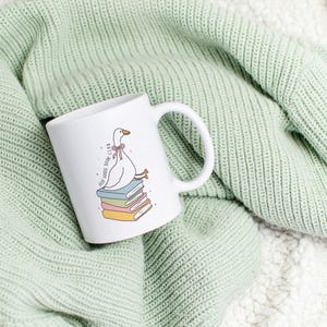May include: White ceramic mug featuring a cartoon goose wearing a pink bow, perched atop a stack of colorful books. The mug has the text "Self-Goose Book Club" and a white handle. The mug is set against a light green knitted blanket.