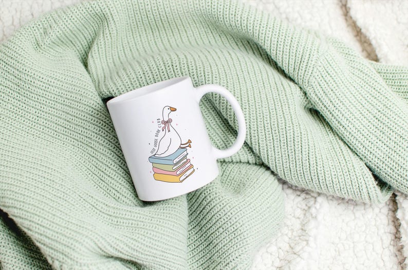 May include: White ceramic mug featuring a cartoon goose wearing a pink bow, perched atop a stack of colorful books. The mug has the text "Self-Goose Book Club" and a white handle. The mug is set against a light green knitted blanket.
