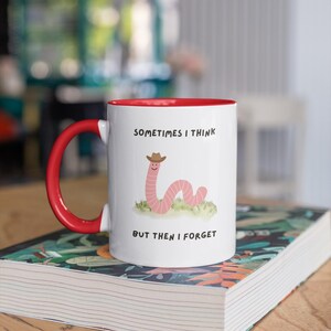 May include: White ceramic mug with a red interior and handle. The mug features a cartoon worm wearing a cowboy hat, with the text "SOMETIMES I THINK BUT THEN I FORGET". The mug is sitting on a book.