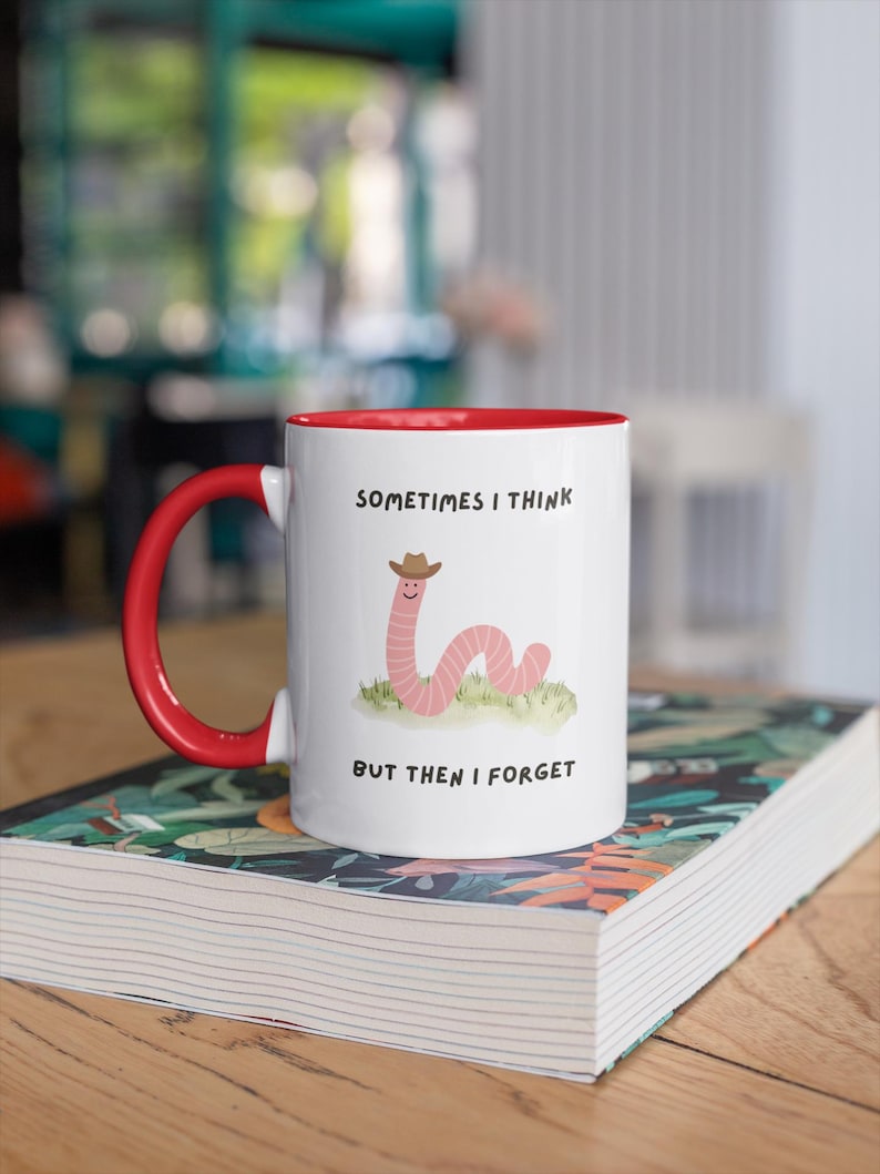 May include: White ceramic mug with a red interior and handle. The mug features a cartoon worm wearing a cowboy hat, with the text "SOMETIMES I THINK BUT THEN I FORGET". The mug is sitting on a book.