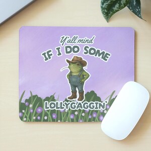 May include: Rectangular mouse mat featuring a cartoon frog in a cowboy hat and dungarees. Text reads "Y'all mind IF I DO SOME LOLLYGAGGIN'". The background is lavender with green grass and purple flowers.