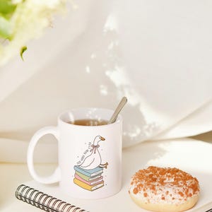 May include: A white ceramic mug with a cartoon goose design, a spoon, a donut, and a notebook. The mug features a goose sitting on books with the text "Self Goose Book Club". The donut has white frosting and a brown topping.