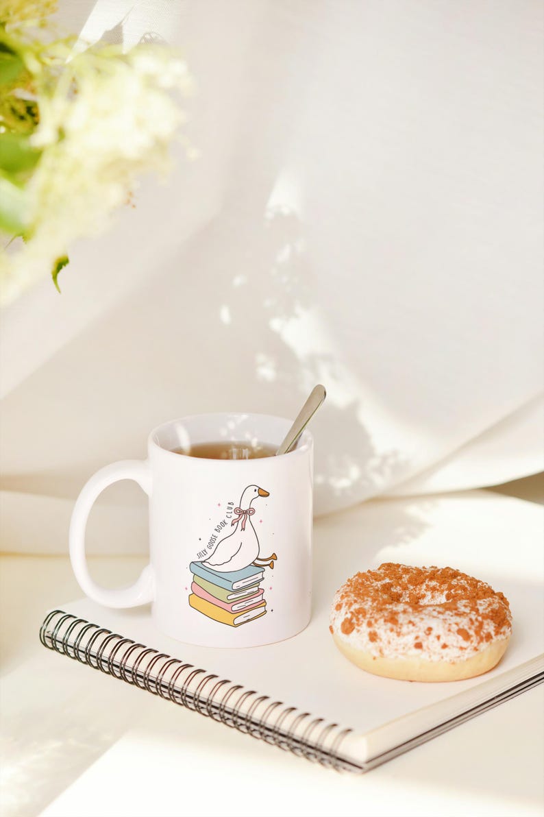 May include: A white ceramic mug with a cartoon goose design, a spoon, a donut, and a notebook. The mug features a goose sitting on books with the text "Self Goose Book Club". The donut has white frosting and a brown topping.