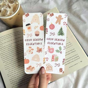 May include: Two rectangular bookmarks with a cozy season theme. One features a sweater, pumpkin spice latte, and fall leaves. The other shows a gingerbread man, Christmas tree, and a mug of hot cocoa. The text "COSY SEASON ESSENTIALS" is printed on both.