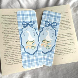 May include: Two blue and white plaid bookmarks with a whimsical design. Each bookmark features a white goose on a stack of books, a teacup, and the text "Just a silly bookish goose."