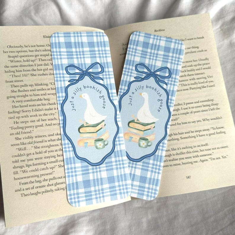 May include: Two blue and white plaid bookmarks with a whimsical design. Each bookmark features a white goose on a stack of books, a teacup, and the text "Just a silly bookish goose."