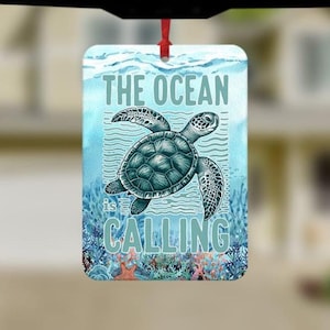May include: A rectangular ornament with a sea turtle design. The ornament features the text "THE OCEAN is CALLING" in a light blue font. The background is a watercolor-style image of the ocean with a turtle swimming in the water. A red ribbon is attached for hanging.