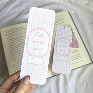 May include: Two pink and white gingham bookmarks with the text "Fell asleep here." One bookmark has a pink tassel. The design includes a pink oval frame, a bow, and an open book illustration.