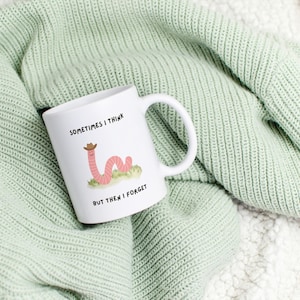 May include: White ceramic mug with a whimsical illustration of a pink worm wearing a cowboy hat, resting on a patch of green grass. The mug has the text "SOMETIMES I THINK BUT THEN I FORGET". The mug is set on a light green knitted blanket.