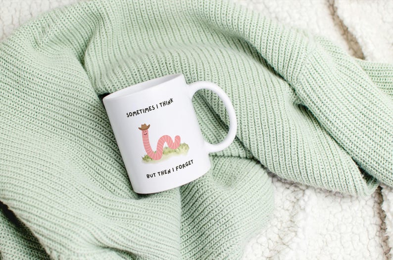 May include: White ceramic mug with a whimsical illustration of a pink worm wearing a cowboy hat, resting on a patch of green grass. The mug has the text "SOMETIMES I THINK BUT THEN I FORGET". The mug is set on a light green knitted blanket.
