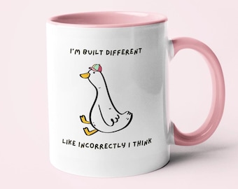 May include: White ceramic mug with a pink interior and handle. The mug features a cartoon duck wearing a hat, with the text "I'm built different, like incorrectly I think."