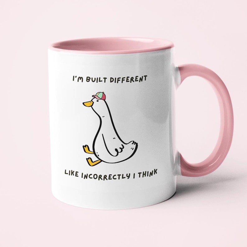Puede incluir: Taza de cer&aacute;mica blanca con interior y asa rosas. La taza presenta un pato de dibujos animados con sombrero, con el texto "I'm built different, like incorrectly I think."